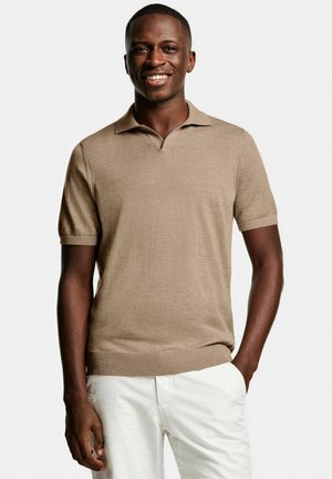 Smiling man wearing a beige short-sleeve polo shirt and white pants, standing with one hand in pocket against a plain background.