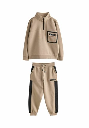 Beige tracksuit-setti mustilla sivupaneeleilla, jossa on puolivetoketjullinen pusero edessä olevalla taskulla ja kiristysnauhalla varustetut jogger-housut suurilla taskuilla.