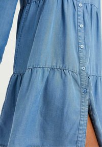 Ljusblå denimklänning med långa ärmar, knäppning fram, samlad midja och sidoflin. Slät textur och subtila sömnadsdetaljer.