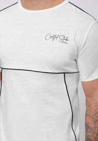 Baltas trumpas rankovėmis marškinėliai iš minkštos medžiagos, su juodomis siūlėmis ir „Crafted Studio 2001“ logotipu juodu raštu.