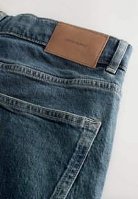 Farkut denim sinisellä pesulla, joissa on ruskea nahkainen takapatch, jossa on teksti "DENIM SUPPLY", sekä klassinen viisi taskua -suunnittelu.