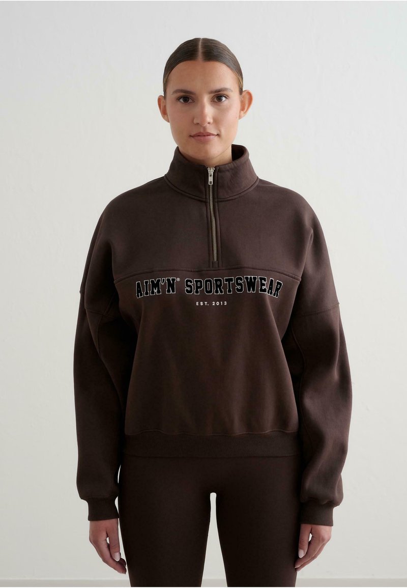 Sudadera marrón con cuello alto y media cremallera. Presenta un destacado texto en blanco "AIM'N SPORTSWEAR EST. 2013" en la parte frontal.