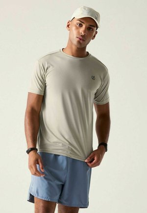 Young man wearing light beige cap and t-shirt with blue shorts, holding t-shirt hem, standing against plain light background.