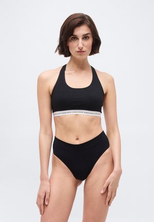 Kvinde iført sort sports-bh med elastikbånd med mærke og højtsiddende sorte trusser, stående mod en ensartet lys baggrund.