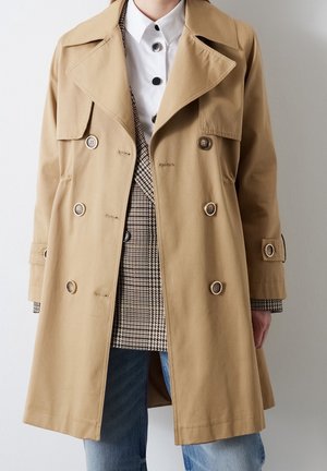 Beige double-breasted trench coat over houndstooth blazer, white shirt with black buttons, and blue jeans on person standing against plain background.
