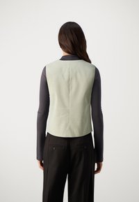 Hellgrünes ärmelloses Top mit glatter Textur, flachen Nähten und tailliertem Design, kombiniert mit einem dunklen Langarmshirt und schwarzen Hosen.