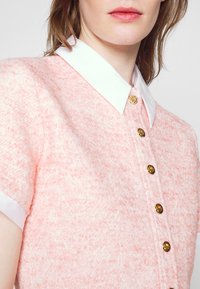 Femme portant un top rose texturé à manches courtes avec un col blanc et des boutons dorés sur le devant, vue des épaules aux lèvres.