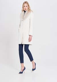 Cappotto bianco con colletto in pelliccia sintetica, tre bottoni, tasche laterali; abbinato a jeans skinny blu scuri e tacchi blu con punte dorate.