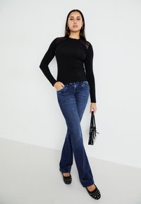 Langärmliges schwarzes Oberteil mit Spitzenakzenten, kombiniert mit blauen Schlagjeans, schwarzen flachen Schuhen mit Nieten und einer kleinen schwarzen Handtasche.