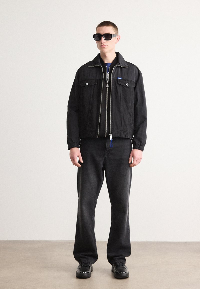 Czarna kurtka zapinana na zamek z dwoma kieszeniami na piersi, zestawiona z ciemnymi dżinsami i czarnymi okularami przeciwsłonecznymi. Posiada elastyczne mankiety i niebieską końcówkę zamka.