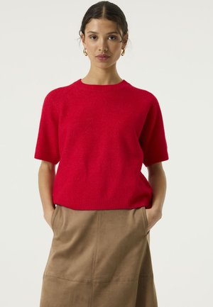 Vrouw staat frontaal, draagt een felrode trui met korte mouwen en een beige rok, handen in de zakken, neutrale achtergrond.