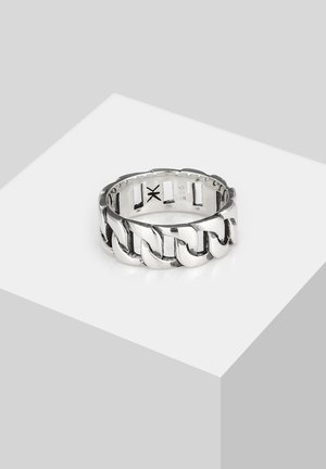 Sølv kæde-link stil ring med indgraverede mærker indvendigt, placeret på en hvid geometrisk piedestal mod en lysegrå baggrund.