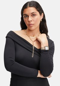 Schwarzes, schulterfreies Oberteil mit langen Ärmeln und drapiertem Design. Akzentuiert mit geschichteten goldenen Halsketten und einer silbernen Uhr. Strukturierter Stoff.