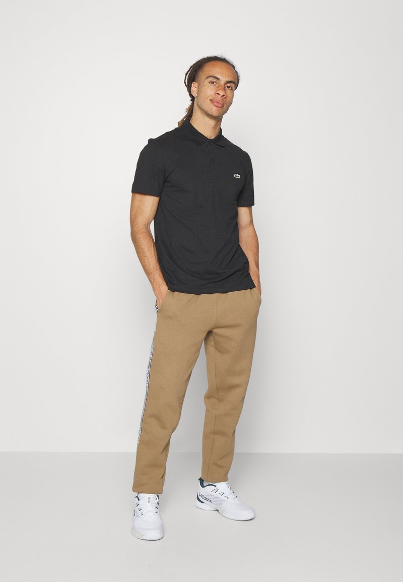Polo noir à manches courtes avec un logo vert, associé à un pantalon de jogging beige et des baskets blanches. Design simple avec une coupe classique.