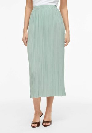 Femme portant une jupe midi plissée vert clair avec une taille élastique, associée à des sandales à talons marron à bout ouvert.