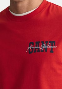 Rotes Baumwoll-T-Shirt mit kurzen Ärmeln, auf der Vorderseite mit schwarzem Text "CANT" und dem Logo "American Sportswear" in Schreibschrift.