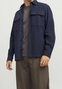 Námořnická vlna košile s kapsami na hrudi, s knoflíkovým uzávěrem a límcem. Nosená přes šedé tričko, v kombinaci s hnědými kalhotami.