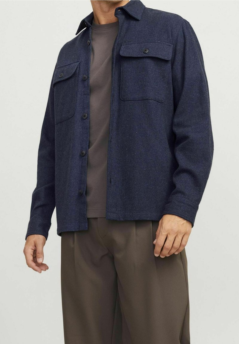 Námořnická vlna košile s kapsami na hrudi, s knoflíkovým uzávěrem a límcem. Nosená přes šedé tričko, v kombinaci s hnědými kalhotami.