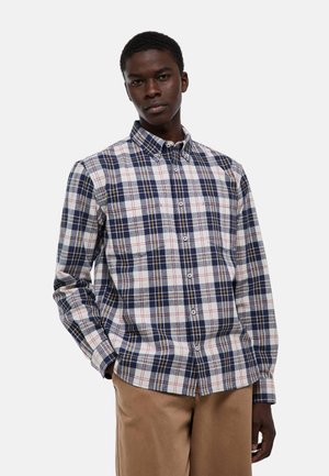 Jeune homme portant une chemise à carreaux bleue, blanche et rouge à boutons, ainsi qu'un pantalon beige, debout avec une main dans la poche.