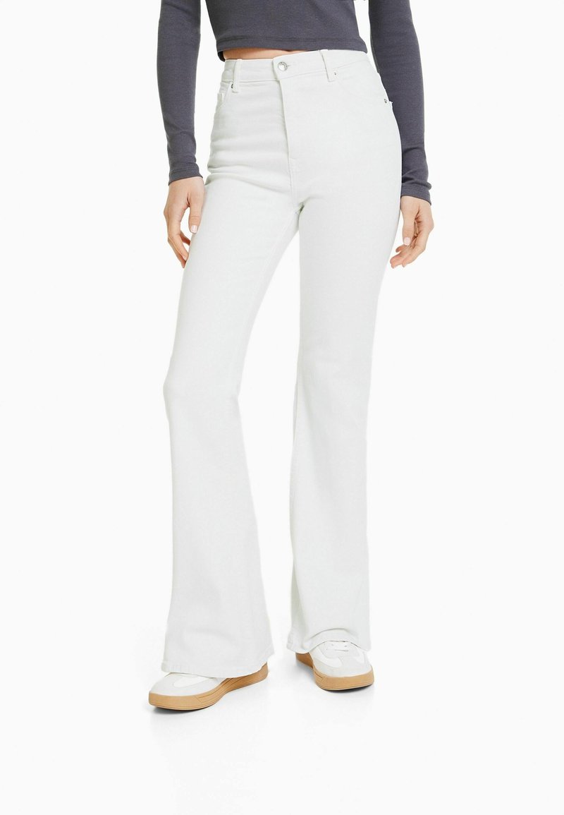 Femme portant un jean évasé blanc taille haute, des baskets blanches et un crop top gris foncé à manches longues, debout les mains détendues.