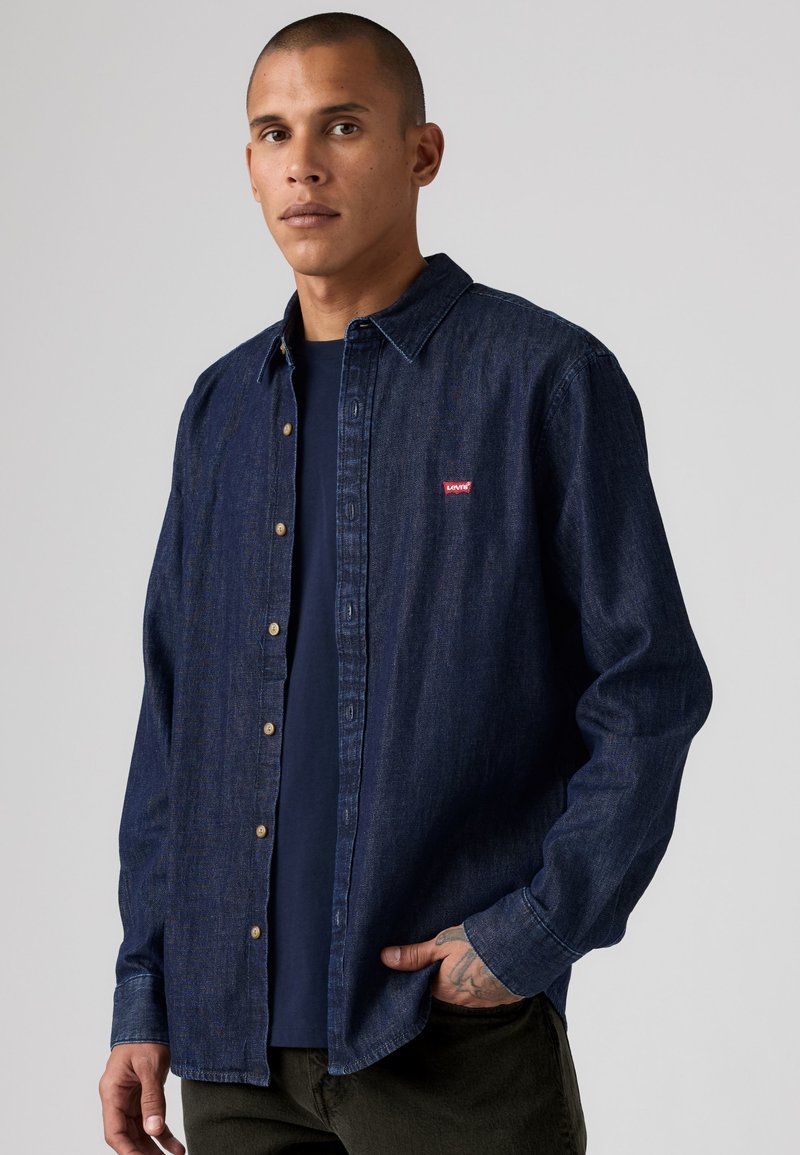 Homme portant une chemise en denim bleu foncé boutonnée sur un t-shirt bleu marine avec une main dans la poche sur un fond gris uni.