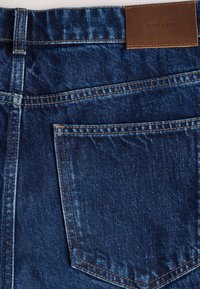 Modré rifľové džínsy v detailnom zábere, zobrazujúce zadný vrecko, pútká na opasok, detaily švov a hnedú koženú značku s nápisom "DENIM SUPPLY".