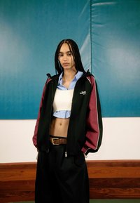 Veste noire et bordeaux avec des accents blancs, chemise boutonnée bleu clair, crop top créme ajusté, pantalon noir à jambes larges, ceinture marron.
