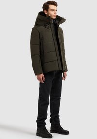 Paminkštintas žalias paltas su aukštu apykakliu ir užtrauktuku, turintis lygią tekstūrą, šonines kišenes ir reguliuojamą gaubtą, derinamas su juodomis džinsais.