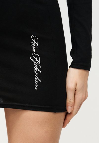 Μαύρη εφαρμοστή φούστα με λευκή κεντημένη καλλιγραφική επιγραφή "Han Høttersleur" στο πλάι, φορεμένη από άτομο με το χέρι να ακουμπάει κοντά στη βάση.