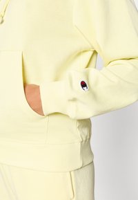 Ανοιχτό κίτρινο φούτερ με κουκούλα από απαλό ύφασμα. Διαθέτει μπροστινή τσέπη τύπου ποδιάς, ριμπ μανσέτες και ένα μικρό λογότυπο στο μανίκι.