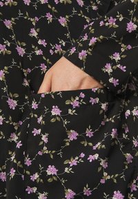 Tela negra con un patrón floral de flores moradas, hojas verdes y acentos beiges, que presenta una mano visible en un bolsillo lateral.