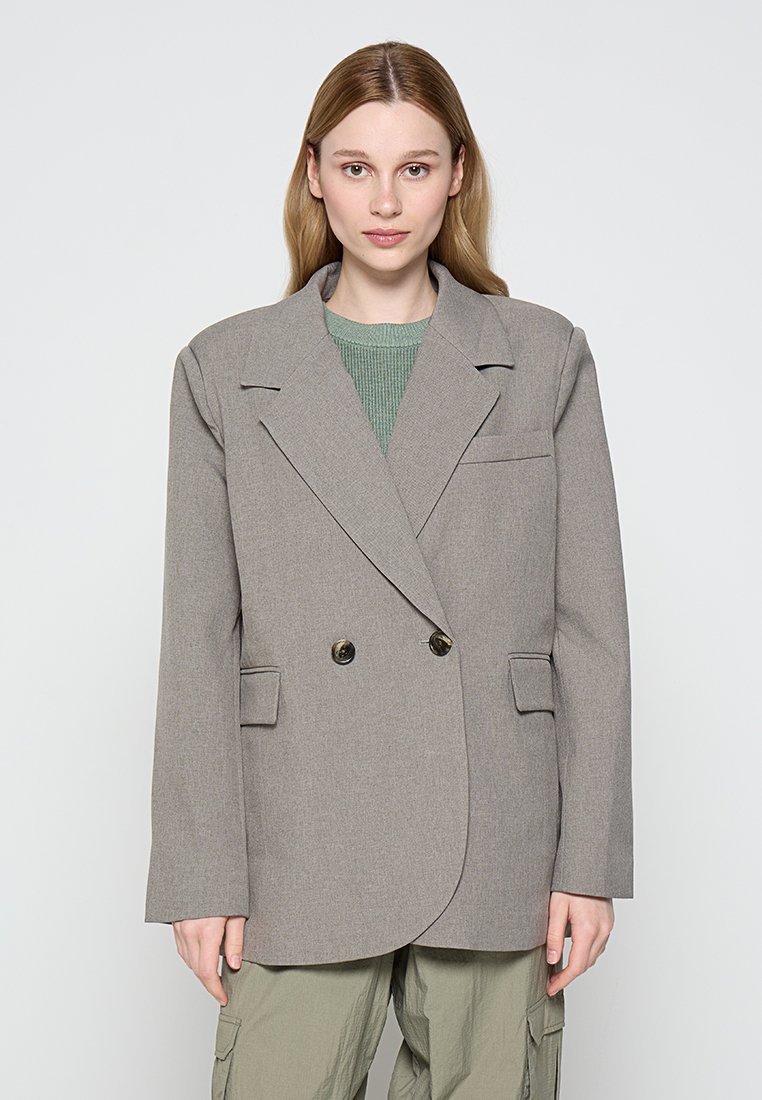 Blazer gris de doble botonadura con solapas en muesca, dos bolsillos frontales y botones decorativos. Llevado sobre una blusa verde de canalé. Textura suave.