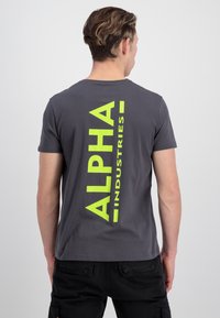 Grå bomulds t-shirt med et stort vertikalt neongrønt "ALPHA" print på ryggen, korte ærmer og en klassisk rund halsudskæring.