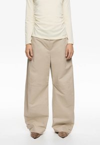 Pantalones de pierna ancha beige hechos de una tela suave, con bolsillos frontales asimétricos y puños con detalles ajustables de hardware.