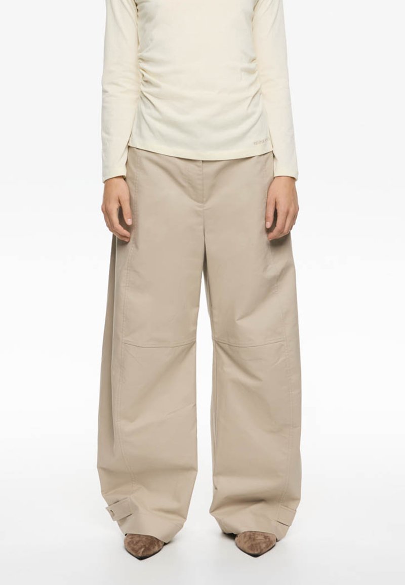 Pantalones de pierna ancha beige hechos de una tela suave, con bolsillos frontales asimétricos y puños con detalles ajustables de hardware.