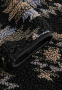 Tissu en polaire noir avec des motifs abstraits beige et bleu. Présente une surface texturée et une bordure en tissu noir le long du bord.