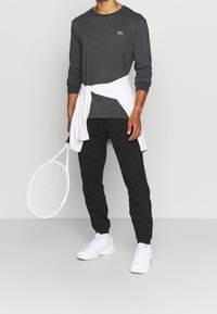 Grå långärmad skjorta, vit slips runt midjan, svarta byxor, vita sneakers, håller en tennisklubba. Enkelt mönster, inga mönster, avslappnad passform.