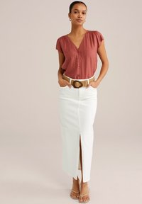 Blusa corta di colore ruggine con bottoni abbinata a una lunga gonna di jeans bianca con uno spacco laterale. Cintura intrecciata beige con fibbia circolare.