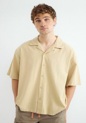 Młody mężczyzna z kręconymi włosami, ubrany w beżową koszulę z krótkim rękawem na guziki i brązowe spodnie, z rękami w kieszeniach, na białym tle.