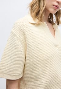 Γυναίκα με πλεκτό πόλο πουκάμισο σε εκρού χρώμα, με κοντά μανίκια, υφή, γιακά και άνοιγμα V στο λαιμό.