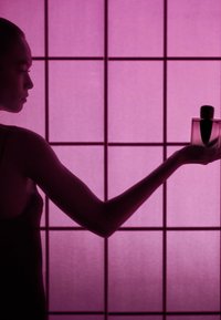 Un frasco de perfume de vidrio claro con una tapa negra, sostenido en una mano. El fondo presenta un panel translúcido rosa con patrones de rejilla.