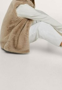 Gilet in finta pelliccia color tan con design senza maniche, caratterizzato da una texture morbida, abbinato a una leggera camicia a maniche lunghe e pantaloni chiari.