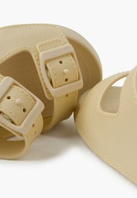 Sandali beige con texture, dotati di due cinturini regolabili con chiusura a fibbia; punta rotonda e sezioni aperte per la traspirabilità.