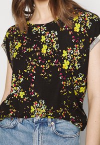 Czarna bluzka z krótkim rękawem w kwiatowy wzór w kolorach żółtym, różowym i zielonym. Miękka tkanina i luźny krój, z szarym akcentem przy rękawach.