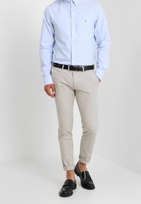 Lichtblauw overhemd met lange mouwen en knopen, beige slim-fit chino's en zwarte loafers. Het overhemd heeft een borstzak aan de linkerkant; de chino's zijn opgerold bij de manchetten.