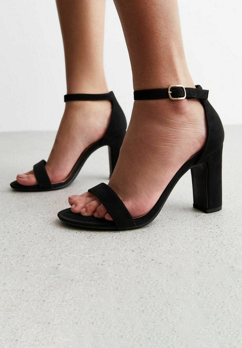 Sandali con tacco alto in suede nero, dotati di una larga fascia alla caviglia e punta quadrata. Presentano una fibbia dorata e una suola testurizzata.