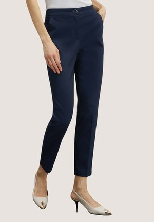 Slim-fit marineblå bukser med sidelommer iført hvide spidse slingback-hæle med metalliske spidser, vist på en stående person.