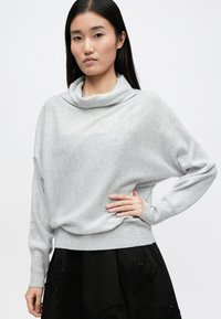 Ανοιχτό γκρι oversized πουλόβερ με λαιμόκοψη, μακριά μανίκια και ριμπ μανσέτες, συνδυασμένο με μαύρη φούστα που έχει κεντημένα σχέδια.