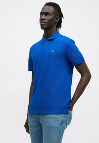 Kék galléros póló rövid ujjakkal és egy kisebb logóval a mellkason. Világoskék farmernadrággal viselve. Simább textúra.