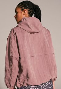 Giacca con cappuccio rosa realizzata in tessuto leggero e testurizzato. Presenta spalle cadenti e polsini elastici. Leggera lucentezza e vestibilità rilassata.
